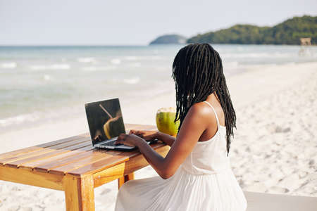 Young Black woman with dreadlocks sitting at wooden table on beach and working on laptopの写真素材