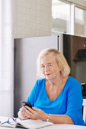 Portrait of smiling elderly woman sitting at desk in kitchen and answering text messages from her...