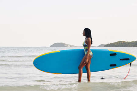 Serious pretty fit young woman carrying sup board and looking at sea wavesの写真素材
