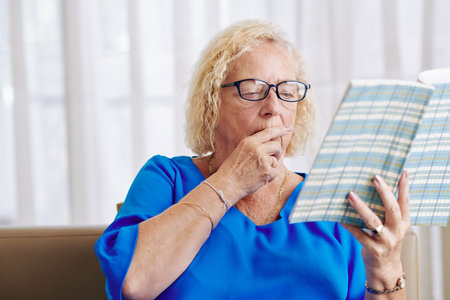 Aged woman in glasses sitting on sofa in living room and reading captivating book