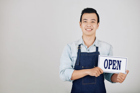 Happy young cafe owner in denim apron holding open sign, isolated on grey