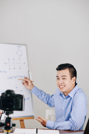 Young smiling chemistry teacher pointing at chemical formula on whiteboard when conducting online...