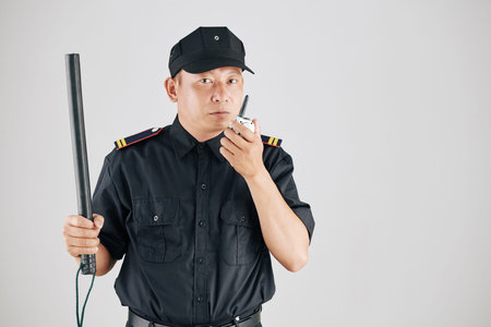 Studio photo of serious police officer raising hand with truncheon and talking to chief of police via walkie-talkieの写真素材