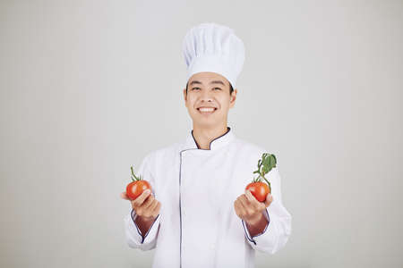 Portrait of happy handsome young chef in uniform holding fresh ripe tomatoesの写真素材