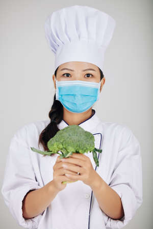 Young female cafe chef in medical mask holding fresh broccoliの写真素材
