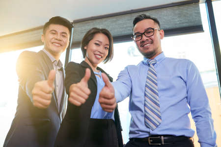 Group of three Asian office workers showing thumb up gesture smiling at camera, horizontal low angle shotの写真素材