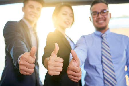 Three modern business people showing thumb up gesture smiling at camera, horizontal back light hands focused shotの写真素材
