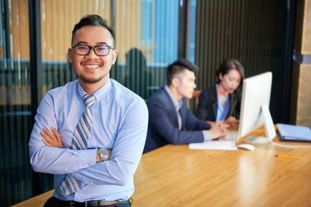 Horizontal medium portrait of confident Asian businessman wearing eyeglasses standing with arms crossed smiling at camera, his colleagues working behind himの写真素材