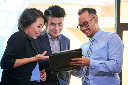 Group of three brand managers standing together working on business plan looking at digital tablet...