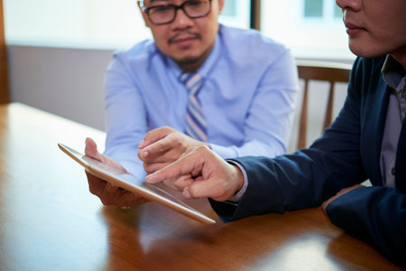 Two Asian male office workers sitting together at table using Internet on digital tablet, horizontal...