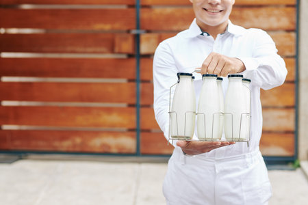 Young milkman in perfect white uniform standing against wooden fence holding glass bottles with...
