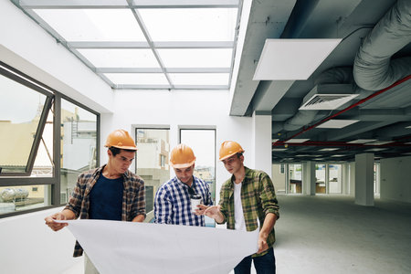 Three Middle Eastern men wearing hardhats standing together in empty room looking at construction...