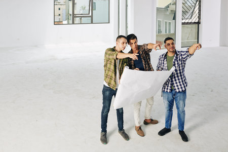 Group of three professional engineers standing in empty spacious room holding building plan paper...