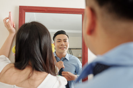 Young Asian couple standing in front of mirror when getting ready in the morning. Man tying necktie and woman applying make-upの写真素材