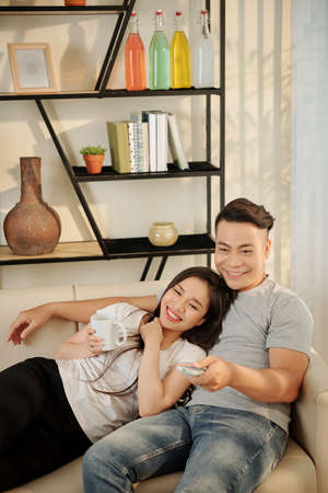 Cheerful young Vietnamese couple resting on sofa at home and watching comedy movieの写真素材