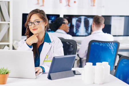 Portrait of pretty smiling young female doctor in glasses sitting at office desk with laptop, tablet...
