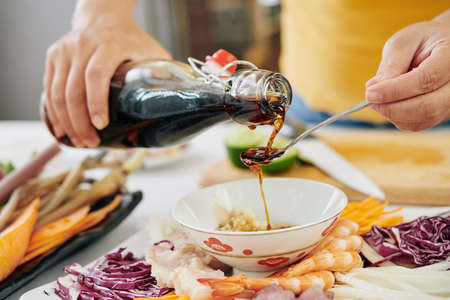 Woman pouring couple of tea spoons of soy sauce into small bowl when making dipping sauce for snack plateの写真素材
