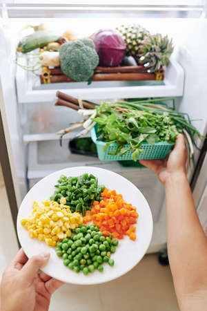 Hands of person opening fridge taking out plate with corn, diced carrot, pea and grean beansの写真素材