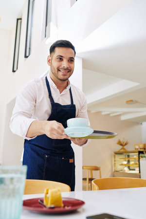 Handsome smiling waiter with beautiful smile bringing cup of cappuccino and piece of cake for...