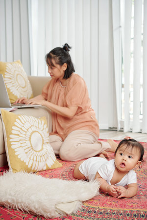 Young woman sitting on the floor and working on laptop when little daughter crawling near by