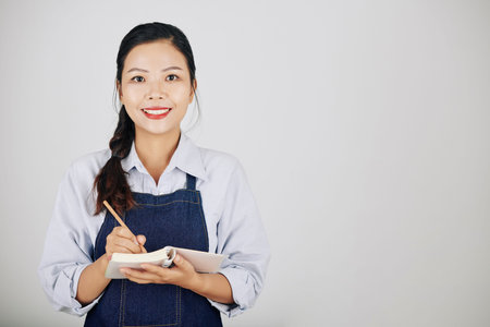 Portrtait of smiling pretty young waitress writing down order of customer