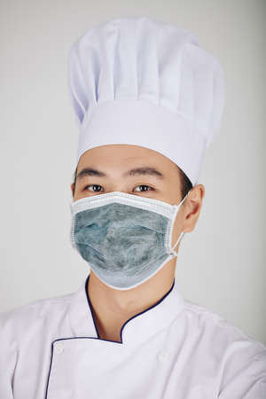 Portrait of young serious Asian restaurant chef in hat and medical mask looking at cameraの写真素材