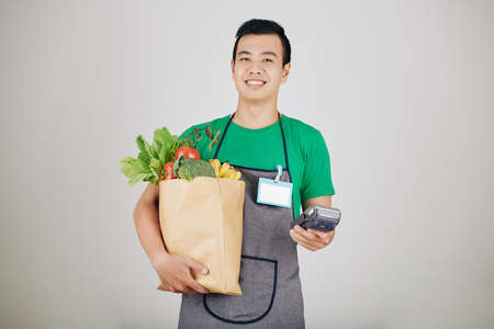 Portrait of smiling young supermarket worker with paper package with goceries and payment terminalの写真素材