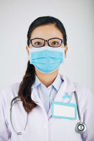 Portrait of young female general practitioner in glasses and medical mask with badge on her labcoatの写真素材