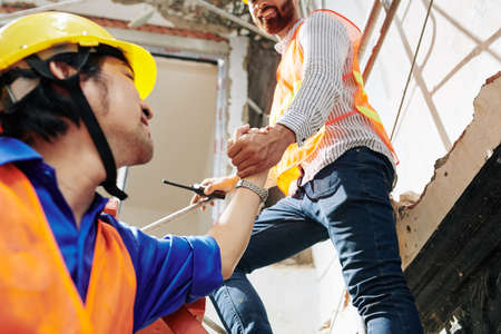 Smiling construction worker outstretching hand when helping colleague to get on ladder next to himの写真素材