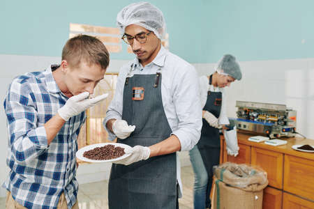Roastery worker letting customer to smell freshly roasted coffee beans, his colleague packing beans in backgroundの写真素材