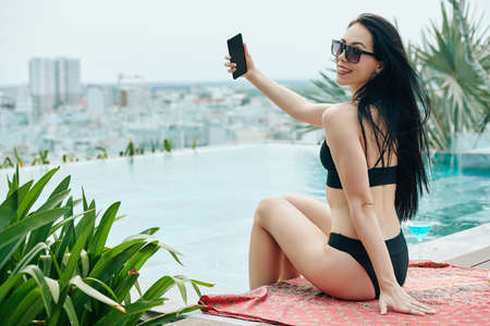 Happy beautiful young woman spending time by infinity pool and taking selfieの写真素材