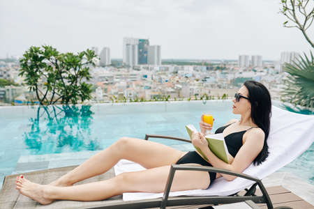 Pretty slim young woman relaxing on chaise-lounge near infinity pool with book and glass of orange juce and looking at big cityの写真素材