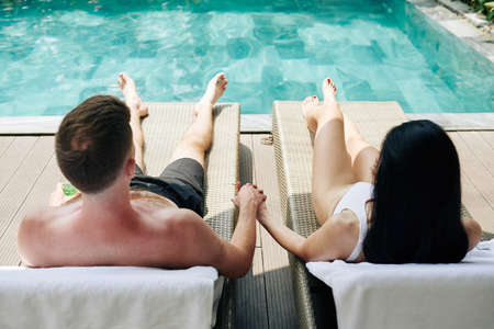 Young couple holding hands when sunbathing by swimming pool and lookin at waterの写真素材