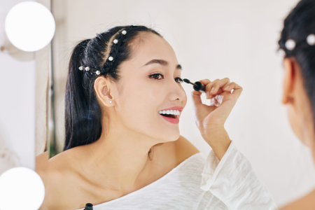Happy pretty young Vietnamese woman applying mascara when getting ready for party at her vanity