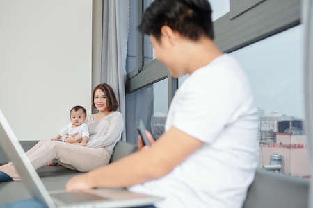 Young woman and her little daughter sitting on sofa and looking at husband working on laptopの写真素材