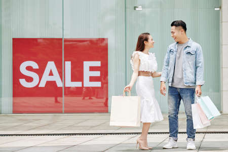 Cheerful young Asian couple standing at big shop window with sale bannerの写真素材