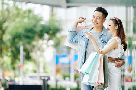 Beautiful happy young Asian couple standing in the street with shopping bags and looking awayの写真素材