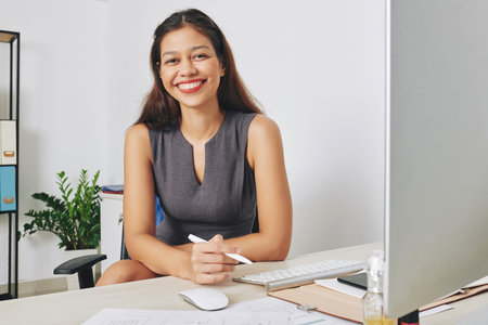 Portrait of young smiling female UI designer working on computer at her desk and sketching project...