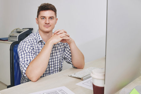 Portrait of handsome smiling young entrepreneur sitting at office desk and looking at camera