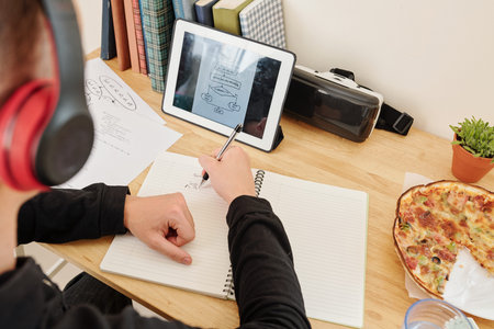 School student sitting at his desk at home, watching programming online course and drawing scheme in...