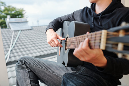 Cropped image of teenager sitting on window sill and enjoying playing guitar