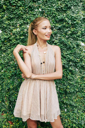 Stylish slim young woman standing at green hedge of fower garden, smiling at looking awayの写真素材