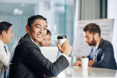 Portrait of smiling mature businessman with cup of take away coffee attending meeting with colleaguesの写真素材