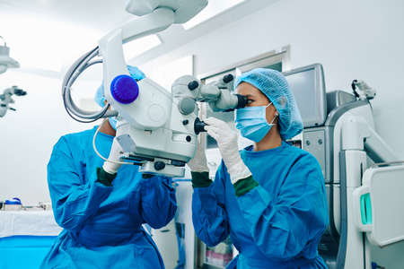 Asian female surgeon in scrubs using electronic microscope when operating patientの写真素材