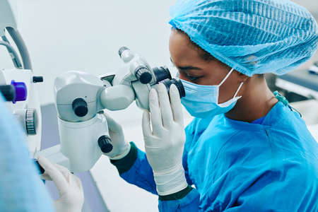 Young Black female surgeon in medical mask and rubber gloves looking through electronic miscroscopeの写真素材
