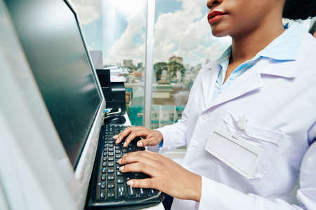 Cropped image of serious young Black female doctor working on computer in clinic and checking documents of patientの写真素材