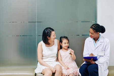 Smiling friendly young Black pediatrician talking to little girl and her motherの写真素材