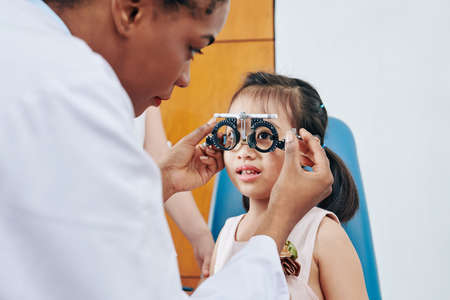 Pediatric optometrist helping little girl to put on test glasses when checking her eyesightの写真素材