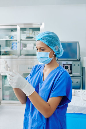 Serious young surgical assistant in scrubs and medical mask putting on sterilized surgical gloves...