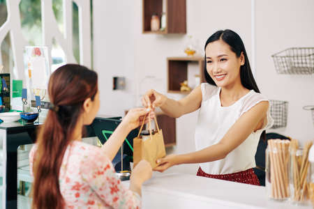 Happy pretty young Vietnamese shop assistant giving paper bag with order to female customerの写真素材
